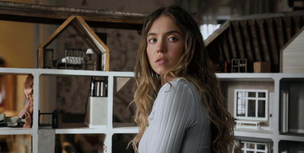 Woman with long wavy hair looks over her shoulder while standing beside a detailed dollhouse model in a dimly lit room.