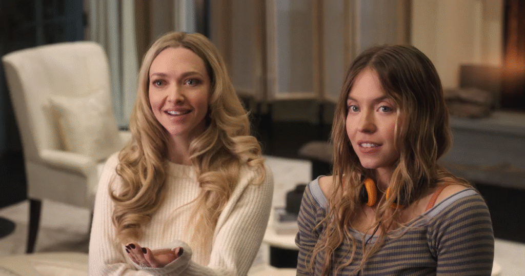 Two women sit indoors on a sofa, smiling toward the camera; one has long blonde curls in a cream sweater, the other has wavy light-brown hair wearing a striped top with orange headphones around her neck.